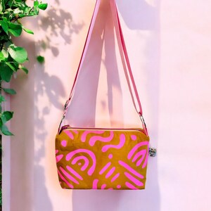 May include: A mustard yellow crossbody bag with a pink abstract pattern. The bag has a pink adjustable strap.