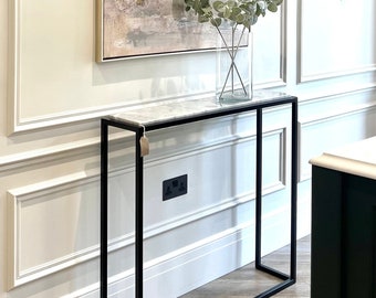 May include: A modern entryway scene featuring a black metal console table with a marble top. A clear glass vase with greenery sits on the table. Above, a large abstract painting in neutral tones with gold accents hangs on the wall.