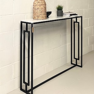 May include: A white marble top console table with a black metal frame. The table has a geometric design and features a rectangular top with a simple, clean design.
