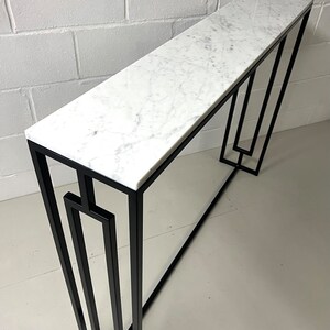 May include: A contemporary console table featuring a white marble top and a black metal frame. The table has a rectangular shape and a geometric design on the legs. The marble top has grey veining.