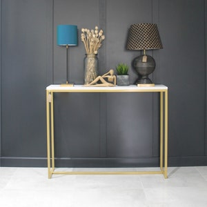 May include: A gold metal console table with a white marble top. The table has a teal blue lamp with a white shade, a black lamp with a gold patterned shade, a small potted plant, and a decorative figurine.
