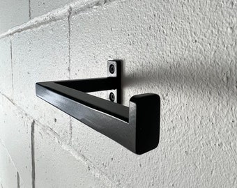 May include: Black metal toilet paper holder mounted on a white brick wall. The holder is angled and has a square end.