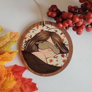 May include: A round wooden ornament with a painted image of a person sleeping with a brown eye mask on a bed with fall leaves. The ornament has a twine string for hanging.