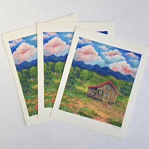 May include: Three prints of a painting depicting a small wooden cabin with a red roof in a green field with blue mountains in the background. The sky is blue with pink clouds.