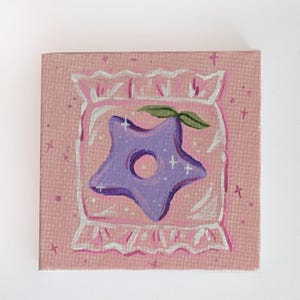 May include: A hand-painted canvas depicting a purple star-shaped cookie with a green leaf, inside a white and pink candy bag with a white lace trim. The background is pink with white dots and crosses.