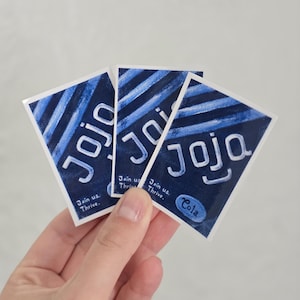 May include: Three blue and white stickers are held in a hand. Each sticker has the word "Jojo" in white, with a blue background and white stripes. The text "Join us. Thrive." is also on the stickers. One sticker says "Cola".