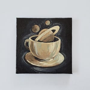 May include: A square painting depicts a teacup and saucer with a planet and stars inside. The teacup is beige, and the planet has a ring around it. The background is black, and the painting is signed "ThePapermegpie".