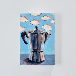 Puede incluir: Pintura original de una cafetera italiana plateada sobre un cielo azul con nubes blancas y naranjas. La pintura está sobre un pequeño lienzo rectangular. La firma del artista, "ThePapermegpie", está en la esquina inferior derecha.