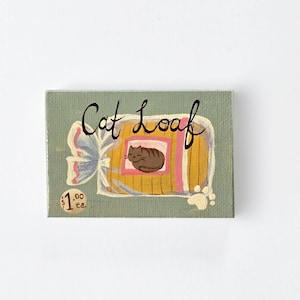 Puede incluir: Un lienzo pintado a mano con un fondo verde claro que presenta una ilustración de dibujos animados de un pan de gato en un paquete amarillo y rosa. El texto "Cat Loaf" está escrito en cursiva negra sobre el paquete. La etiqueta de precio dice "1,00 € cada uno."