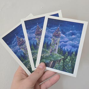 May include: Three art prints depicting a fairytale tower scene. The tower has a purple roof and is surrounded by trees and clouds under a starry night sky. The prints are held in a hand against a white background.