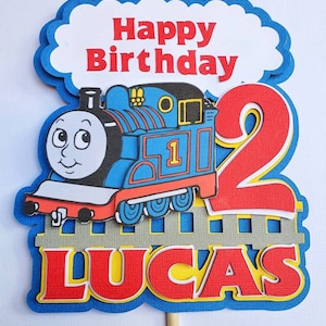 May include: A blue and red Thomas the Tank Engine cake topper with a white cloud that says "Happy Birthday" and a red number 2. The topper also has a grey fence and the name "Lucas" in red letters.