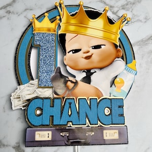 May include: A blue and gold cake topper featuring a cartoon baby wearing a gold crown and a suit, holding a bottle and money. The topper says "CHANCE" in blue letters.