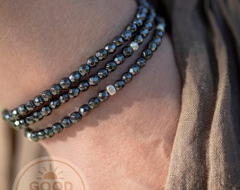 May include: A triple-strand bracelet featuring small, faceted, metallic dark gray beads. The design includes a single, smooth, silver-toned accent bead on each strand. The bracelet is worn on a wrist, with a Good Vibes Sanctuary logo visible.