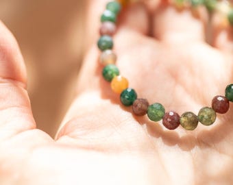 May include: A beaded bracelet with a variety of colors, including green, orange, and maroon. The bracelet is made of small, round beads and is resting on a person's open palm. The beads have a faceted appearance.