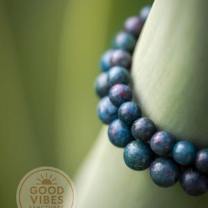 May include: A beaded bracelet with blue and purple spherical beads. The bracelet is wrapped around a green surface. The image includes a circular logo with the text "GOOD VIBES SANCTUARY".