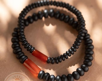 May include: Two black beaded bracelets with a red-orange accent bead. The bracelets are stacked on top of each other. The bracelets are on a tan surface. The image includes the text "GOOD VIBES SANCTUARY".