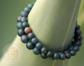 May include: A beaded bracelet with dark green and reddish-brown beads. The bracelet is resting on a light green surface. The beads are round and have a matte finish. The bracelet is a piece of jewelry.