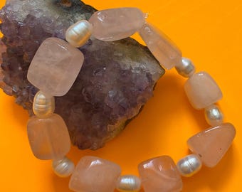 May include: A bracelet made with light pink rose quartz and white pearl beads. The bracelet is on a purple amethyst crystal.