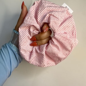 Könnte beinhalten: Ein rosa Haargummi mit einem sich wiederholenden Muster aus kleinen roten Punkten. Das Haargummi ist aus weichem Stoff gefertigt und wird in einer Hand gehalten. Ein kleines weißes Etikett ist am Haargummi befestigt. Dieses Haaraccessoire ist perfekt.