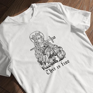 May include: White t-shirt with a black and white illustration of a woman with a crown and a sword through her head. The text "This is fine" is printed below the illustration.