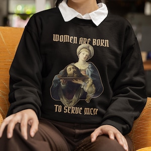 May include: Black sweatshirt with the text "WOMEN ARE BORN TO SERVE MEN" in a vintage font. The graphic depicts a Renaissance-style painting of a woman holding a platter with a head on it. The model is wearing a white collared shirt.