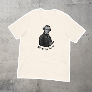 Puede incluir: Una camiseta blanca con una imagen en blanco y negro de un hombre con peluca empolvada y el texto "Serving Kant" impreso en la parte delantera.