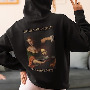 Puede incluir: Sudadera con capucha negra con una pintura de dos personas y el texto "Women are born to serve men".
