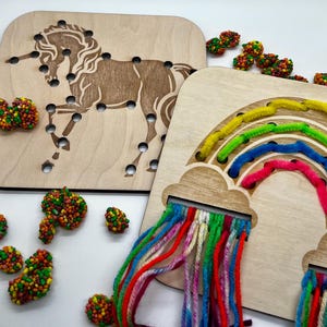May include: Two wooden lacing boards, one with a unicorn design and the other with a rainbow. The unicorn board has a laser-cut design with holes for lacing. The rainbow board features a cloud design with colorful yarn strands. Colorful beads are scattered around the boards.