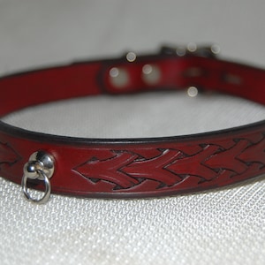 Puede incluir: Un collar de cuero rojo con un diseño trenzado y un anillo de plata.