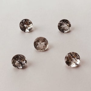 May include: Five clear, oval-shaped gemstones with black inclusions. The stones are arranged in a semi-circle on a white background.