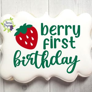 Puede incluir: Una galleta blanca, con bordes festoneados, con un gráfico de fresa roja y las palabras "berry first birthday" en verde. Un pequeño diseño floral está en la esquina superior izquierda. La galleta está sobre una superficie de madera clara.