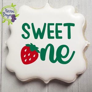 Puede incluir: Una galleta blanca de forma única con texto verde que dice "SWEET one" y una ilustración de fresa roja. La galleta está decorada con un logotipo floral verde y morado en la esquina superior izquierda.