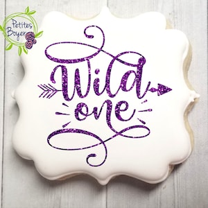 May include: A white cookie decorated with purple glitter lettering that reads "Wild One" with arrows on either side of the text.