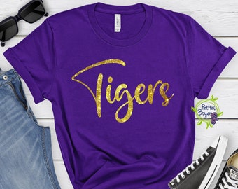 Puede incluir: Una camiseta morada de manga corta con la palabra Tigers en una fuente dorada con purpurina en el pecho. La prenda se muestra junto a unos vaqueros azules, gafas de sol negras y zapatillas de lona bajas en blanco y negro.
