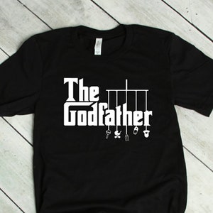 May include: Black t-shirt with white text that reads "The Godfather" with a line of hanging baby toys below the text.