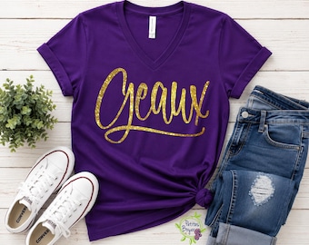 May include: A purple V-neck t-shirt with short sleeves and a rolled cuff detail. The front features the word Geaux written in a gold glitter script font. The shirt is displayed alongside a pair of blue denim jeans and white canvas sneakers.