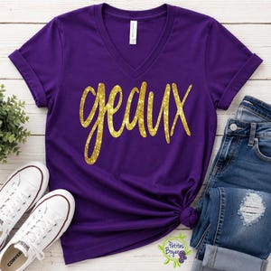 May include: A purple V-neck t-shirt with short sleeves and rolled cuffs. The front features the word geaux in a gold glitter script font. The shirt is displayed alongside blue denim jeans and a pair of white low-top canvas sneakers.
