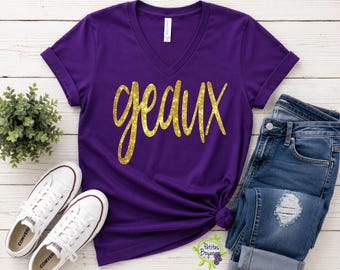 Puede incluir: Una camiseta morada de cuello en V con mangas cortas y puños enrollados. La parte delantera presenta la palabra geaux en una fuente cursiva de purpurina dorada. La camiseta se muestra junto a unos vaqueros azules y zapatillas blancas.