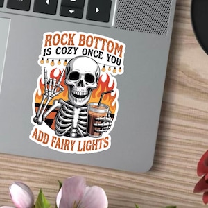 Könnte beinhalten: Aufkleber mit einem Skelett, das ein Kaffeegetränk hält, umgeben von Flammen und Lichterketten. Der Text lautet: "Rock Bottom is Cozy Once You Add Fairy Lights."