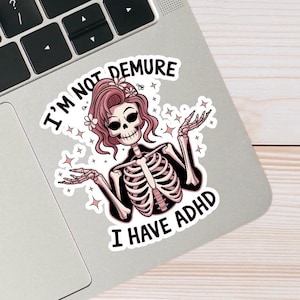 Könnte beinhalten: Ein Aufkleber mit einem Skelett mit rosa Haaren und Blumen, die Arme ausgestreckt. Der Text lautet "I'M NOT DEMURE I HAVE ADHD". Das Design umfasst rosa Stern-Akzente und befindet sich auf einem Laptop.