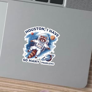 May include: A sticker depicting a raccoon astronaut in a white spacesuit, floating in space. The sticker includes the text "Houston, I have so many problems", along with illustrations of planets and stars.