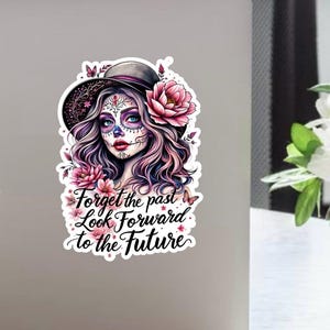Könnte beinhalten: Ein Aufkleber mit einer Sugar-Skull-Frau mit Hut, Blumen und Schmetterlingen. Die Frau hat aufwendiges Gesichtsschminke und lila Haare. Der Aufkleber enthält den Text "Forget the past, Look Forward to the Future."