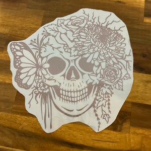Pu&ograve; includere: Un adesivo in vinile bianco con un contorno marrone di uno scheletro con fiori e una farfalla. Lo scheletro ha un grande fiore in cima alla testa e una farfalla sul lato.