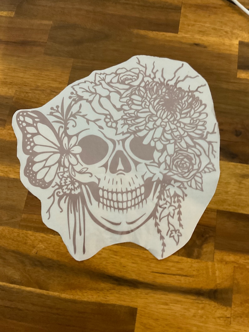 Pu&ograve; includere: Un adesivo in vinile bianco con un contorno marrone di uno scheletro con fiori e una farfalla. Lo scheletro ha un grande fiore in cima alla testa e una farfalla sul lato.