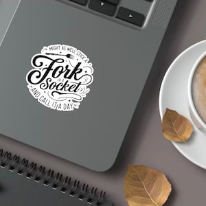 Puede incluir: Una pegatina blanca con el texto "Might as well stick a Fork in a Socket and call it a day" está en un portátil. Una taza de café con arte latte y hojas de otoño están sobre una superficie gris.