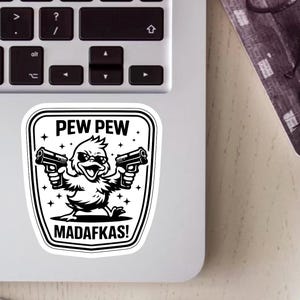 May include: A black and white sticker showing a cartoon duck with sunglasses and guns. The text "PEW PEW" is above the duck, and "MADAFAKAS!" is below. The sticker is on a laptop.