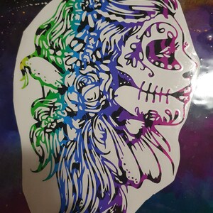 Pu&ograve; includere: Un adesivo colorato di un viso di donna con un design di teschio di zucchero. Il teschio &egrave; delineato in nero e riempito con un arcobaleno di colori. I capelli della donna sono lunghi e fluenti, e ha dei fiori nei capelli.