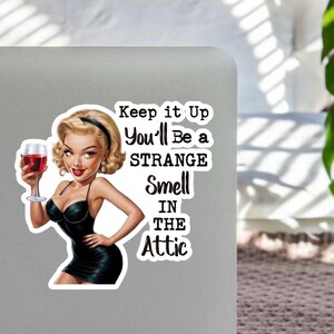 Könnte beinhalten: Aufkleber mit einer Cartoon-Frau in einem schwarzen Kleid, die ein Glas Rotwein hält. Der Aufkleber hat den Text: "Keep it Up, You'll Be a Strange Smell in the Attic."