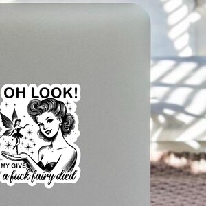 May include: A black and white sticker with a retro illustration of a woman and a fairy. The text "OH LOOK! MY GIVE a fuck fairy died" is part of the design. Suitable for laptops, notebooks, or other personal belongings.