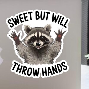 Könnte beinhalten: Aufkleber mit einer Cartoon-Waschbär-Illustration. Der Waschbär wird mit erhobenen Armen dargestellt, und der Text "SWEET BUT WILL THROW HANDS" steht über und unter dem Waschbären. Der Waschbär hat eine schwarze Maske und einen lächelnden Ausdruck.